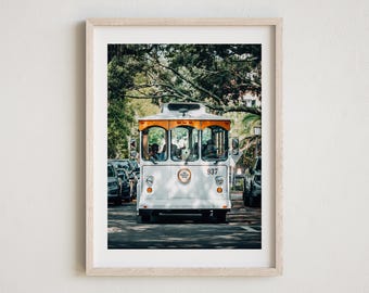 May include: A white trolley with an orange roof and the number 937 on the side, driving down a street lined with trees. The trolley is full of passengers and is surrounded by other cars.