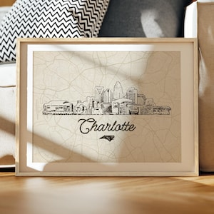 May include: Framed art print featuring a black and white line drawing of the Charlotte, North Carolina skyline. The print includes the city's name and a silhouette of the state. The artwork is displayed in a light-colored wooden frame.