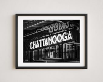 May include: Black and white photograph of a vintage sign that reads "Chattanooga" in white letters on a black background. The sign is attached to a metal structure and is illuminated by lights. The sign is in front of a building with windows.