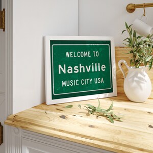 Może przedstawiać: Biały obraz w ramie z zielonym znakiem, na kt&oacute;rym widnieje napis "Welcome to Nashville Music City USA". Obraz leży na drewnianym blacie z białym dzbanem i zieloną rośliną.