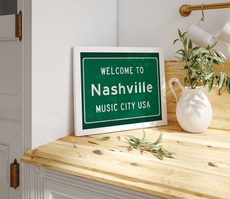 Może przedstawiać: Biały obraz w ramie z zielonym znakiem, na kt&oacute;rym widnieje napis "Welcome to Nashville Music City USA". Obraz leży na drewnianym blacie z białym dzbanem i zieloną rośliną.