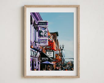 May include: A framed print of a street scene in Nashville, Tennessee. The image features buildings with neon signs, including "World Famous" and "Tootsie's." A guitar is visible on a sign. The sky is blue, and people are walking on the sidewalk.