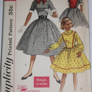 May include: A Simplicity sewing pattern envelope 2203, size 7, featuring illustrations of two dresses. One is a black and white gingham dress with a red collar accent, and the other is a yellow dress with a small red floral print and long sleeves.