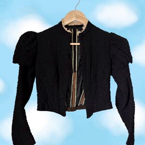 Könnte beinhalten: Eine schwarze Langarmjacke mit einer strukturierten, geknitterten Stoffoberfläche. Das Kleidungsstück verfügt über Puffärmel, einen mit beiger Spitze verzierten Stehkragen und eine Vorderöffnung, die ein dunkles Futter mit vertikalen Streifen zeigt.