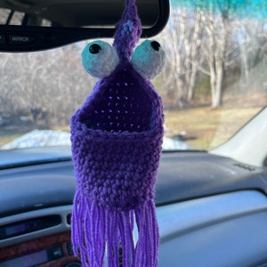 May include: Purple crocheted monster car charm with large white eyes and a beaded mouth. The monster has long purple fringe hanging from the bottom.
