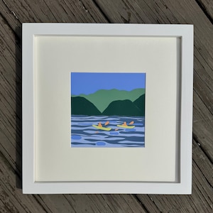 May include: A framed paper art piece depicting a blue lake with green mountains in the background. Three yellow kayaks with orange figures are paddling on the water.