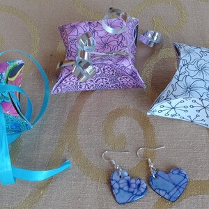 May include: An assortment of gift boxes and heart-shaped earrings. The boxes are decorated with floral patterns and tied with ribbons. The earrings are blue with a marbled design. The items are arranged on a gold-colored surface.