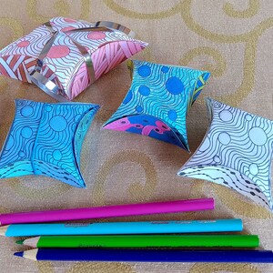 May include: Four colorful paper boxes with geometric patterns, folded into a pillow shape. The boxes are on a beige surface with a patterned design. Four colored pencils are lying on the surface.