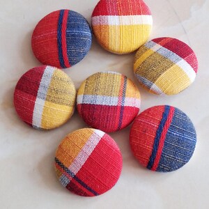 May include: Seven round fabric buttons in various patterns and colors. The buttons feature red, yellow, and blue woven fabric designs. Some have stripes, while others have plaid patterns. The buttons are arranged in a circular pattern.