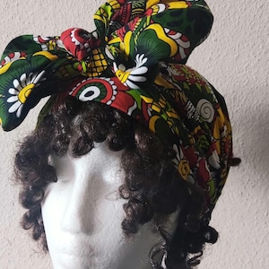 May include: A colorful head wrap with a vibrant floral pattern in red, yellow, green, and white. The head wrap is tied in a bow on top of a mannequin head with dark curly hair. The background is a plain white wall.