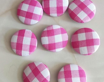 Puede incluir: Ocho botones redondos de tela con un patrón de cuadros vichy rosa y blanco. Los botones están dispuestos sobre una superficie clara. Estos botones son ideales para proyectos de costura y manualidades, añadiendo un toque clásico.