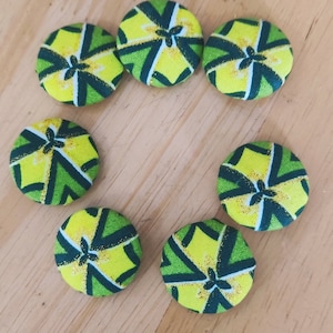 May include: Seven green and yellow fabric buttons with a geometric pattern. The buttons are arranged in a circle on a wooden surface.