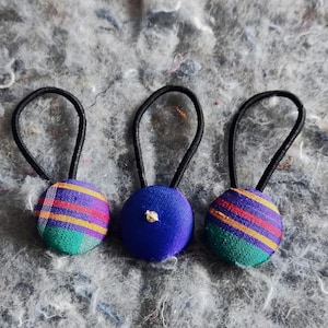 May include: Three hair scrunchies with fabric covered buttons. One scrunchie has a blue button, and the other two have a multi-colored striped button.