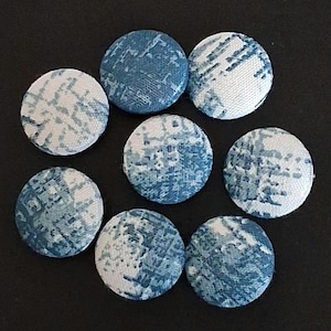 Puede incluir: Ocho botones redondos cubiertos de tela en tonos azules y blancos. Los botones tienen un patrón texturizado y están dispuestos sobre una superficie negra. Estos botones se pueden utilizar para proyectos de costura o manualidades.