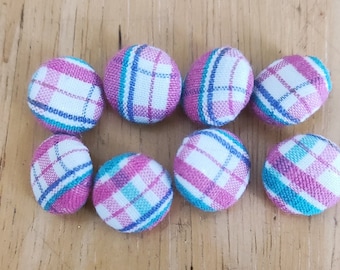 Puede incluir: Diez botones cubiertos de tela, de forma ovalada, con un diseño de cuadros blancos, rosas y azules. Los botones están dispuestos sobre una superficie de madera de color marrón claro. Adecuado para costura.