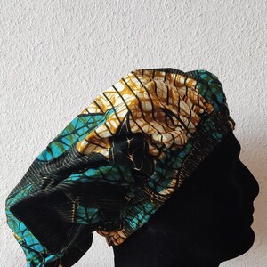 May include: A patterned head wrap in shades of teal, gold, and black, displayed on a black mannequin head. The fabric features a geometric design. The head wrap is a stylish accessory.