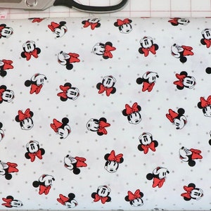 Puede incluir: Tela blanca con un patrón repetido de cabezas de Minnie Mouse rojas y negras con lazos rojos. La tela se mide con una cinta métrica rosa.