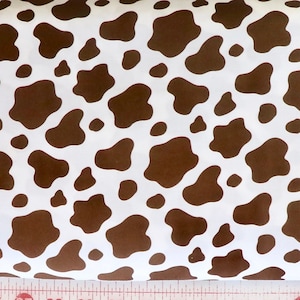 May include: White fabric with a brown cow print pattern. A pink and white measuring tape shows the fabric is approximately 30 cm wide. The fabric has a repeating pattern of irregular brown shapes.
