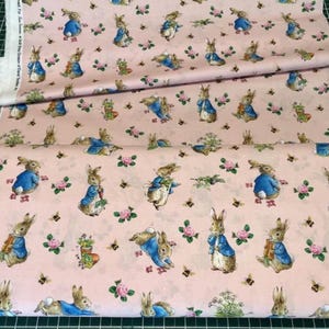 May include: A light pink fabric featuring a repeating pattern of Peter Rabbit characters wearing blue jackets, interspersed with small pink roses, green foliage, and tiny bees. The selvage edge includes printed text identifying the manufacturer.
