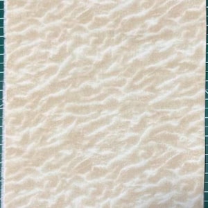 May include: A rectangular piece of fabric with a beige and off-white wavy pattern. The fabric has a soft, textured appearance, resembling sand or a natural landscape. The fabric is placed on a green cutting mat with grid lines.