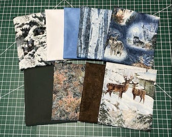 May include: A selection of fabric swatches with nature-inspired patterns. Designs include wolves, deer, snowy scenes, birch trees, and solid colours such as white, blue, and dark green. Suitable for quilting or craft projects.
