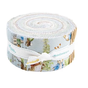 May include: A rolled fabric bundle with a light blue and white pattern, depicting rabbits, butterflies, and text. The fabric is bound with a white ribbon, displaying "BlakeDesigns" and "be colorful".