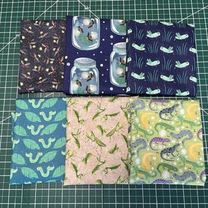 May include: A selection of folded fabric squares featuring insect and reptile designs. Patterns include fireflies, jars, grasshoppers, caterpillars, mantises, and lizards. The fabrics are in shades of blue, green, tan, and black.
