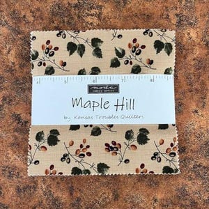 Puede incluir: Una pila de piezas de tela cuadradas con fondo beige y un patrón repetitivo de hojas verde oscuro y pequeñas bayas marrones. Una banda de papel blanco envuelve el centro con el texto "moda", "Maple Hill" y "by Kansas Troubles Quilters", junto a una regla impresa que muestra medidas de hasta 20 cm.