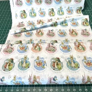 Pode incluir: Um tecido branco com um padrão repetitivo de ilustrações circulares representando personagens de Peter Rabbit. Cada círculo contém o nome de um personagem como Benjamin Bunny ou Cotton-Tail. A estampa inclui cores pastel suaves.