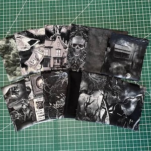 May include: A collection of fabric squares featuring various black and white gothic-themed prints, including images of a skull, a haunted house, a forest, spiders, and tombstones, arranged in a fan-like display on a green cutting mat surface.