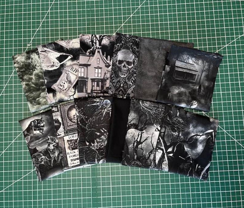 May include: A collection of fabric squares featuring various black and white gothic-themed prints, including images of a skull, a haunted house, a forest, spiders, and tombstones, arranged in a fan-like display on a green cutting mat surface.