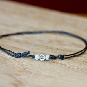 May include: A black string bracelet with two white beads and a silver charm in the center.