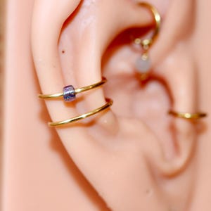 May include: Close-up of an ear adorned with multiple gold-colored hoop earrings. One earring features a small, cylindrical, purple bead. The earrings are arranged in a layered style, creating a stylish and modern look.