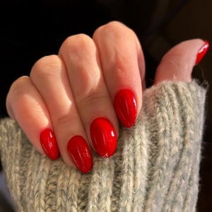 Pode incluir: Mão com verniz vermelho. As unhas são ovais e brilhantes. A mão está parcialmente coberta por um suéter de malha verde claro. O fundo é escuro.