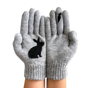 Op de afbeelding: Een paar grijze gebreide handschoenen met zwarte silhouetten van een konijn en een vogel. De handschoenen zijn dubbelgevouwen met de vingertoppen naar de camera gericht.