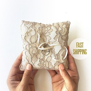 May include: A white lace ring pillow with two gold rings tied with a white ribbon. The pillow is being held by two hands.