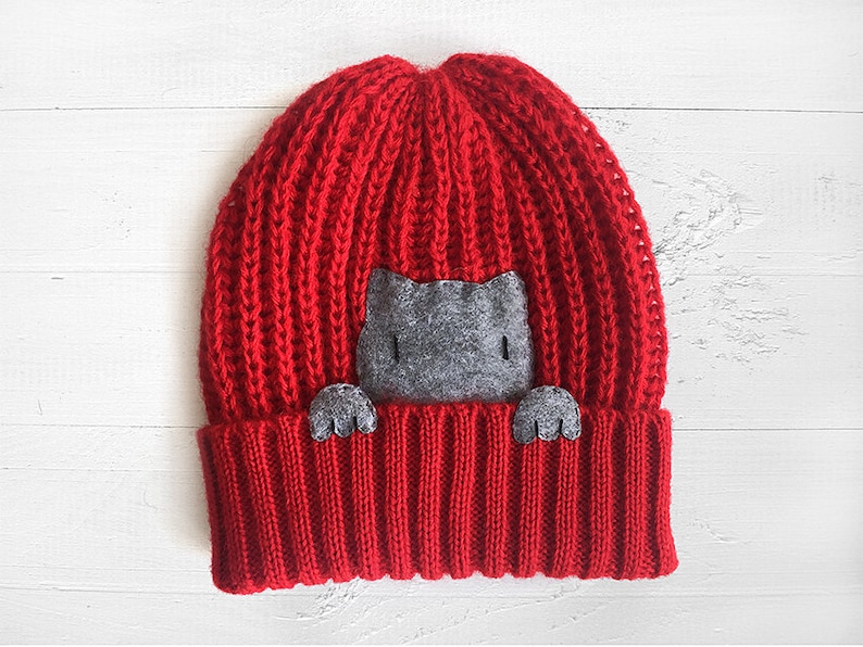 Puede incluir: Gorro de punto rojo con un gato de fieltro gris asom&aacute;ndose por la parte superior.