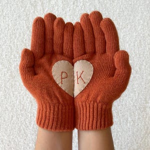 May include: Orange knitted gloves with a beige heart-shaped patch embroidered with the letters "P" and "K" in red. The gloves are shown with the palms facing upwards, showcasing the design.