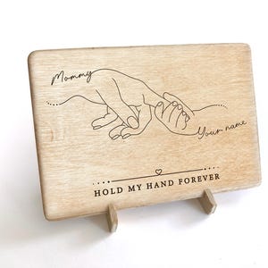 May include: A wooden sign with a hand-drawn design of two hands holding each other. The text "Mommy" and "Your name" are engraved on the sign. The sign also says "Hold my hand forever".