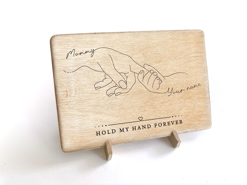 May include: A wooden sign with a hand-drawn design of two hands holding each other. The text "Mommy" and "Your name" are engraved on the sign. The sign also says "Hold my hand forever".