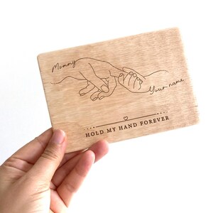 May include: Wooden plaque with a line drawing of two hands holding each other. The text "Mommy" and "Your name" are written above the hands. The text "Hold my hand forever" is written below the hands.