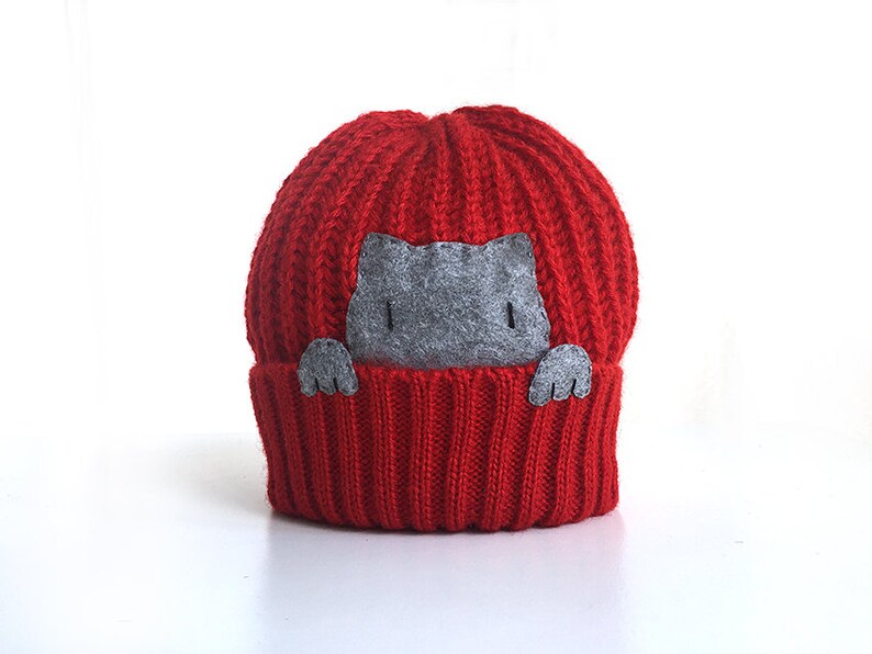 Puede incluir: Gorro de punto rojo con un gato de fieltro gris asom&aacute;ndose por la parte superior.