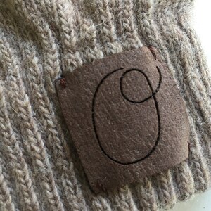 Puede incluir: Un gorro de punto marr&oacute;n con un parche de cuero marr&oacute;n que tiene la letra "O" cosida.
