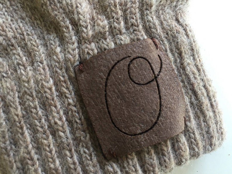 Puede incluir: Un gorro de punto marr&oacute;n con un parche de cuero marr&oacute;n que tiene la letra "O" cosida.