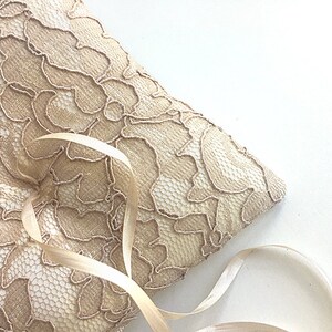 May include: A beige lace ring pillow with a satin ribbon tied around it. The pillow is perfect for holding wedding rings during a ceremony.