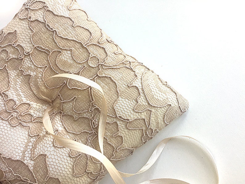 May include: A beige lace ring pillow with a satin ribbon tied around it. The pillow is perfect for holding wedding rings during a ceremony.