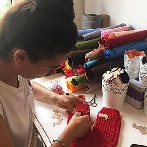 Puede incluir: Una persona est&aacute; cosiendo un parche de fieltro marr&oacute;n en un calientamanos de punto rojo. Hay rollos de fieltro de varios colores en el fondo.