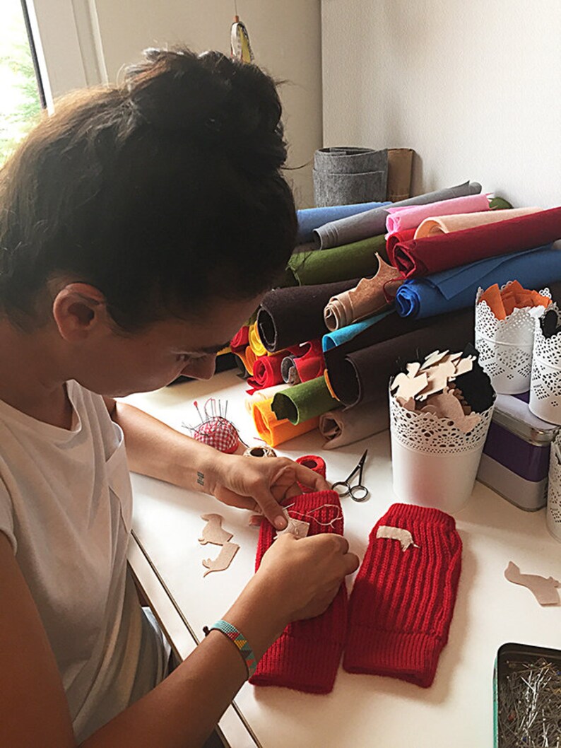 Puede incluir: Una persona est&aacute; cosiendo un parche de fieltro marr&oacute;n en un calientamanos de punto rojo. Hay rollos de fieltro de varios colores en el fondo.