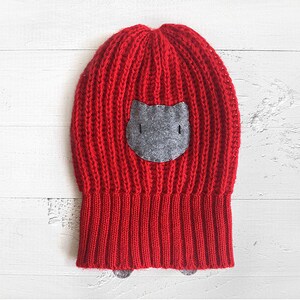 Puede incluir: Gorro de punto rojo con una aplicaci&oacute;n de cara de gato de fieltro gris.
