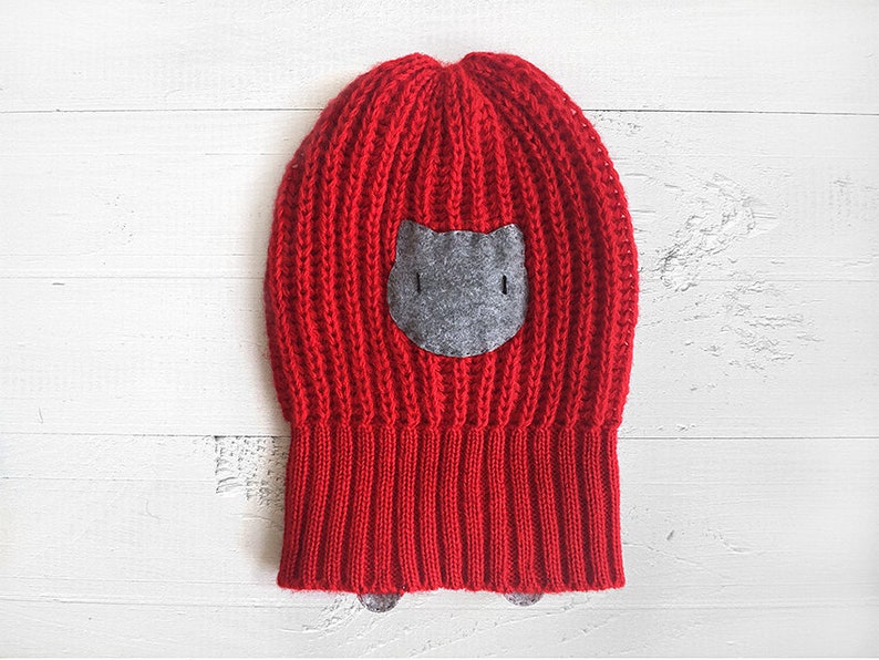 Puede incluir: Gorro de punto rojo con una aplicaci&oacute;n de cara de gato de fieltro gris.